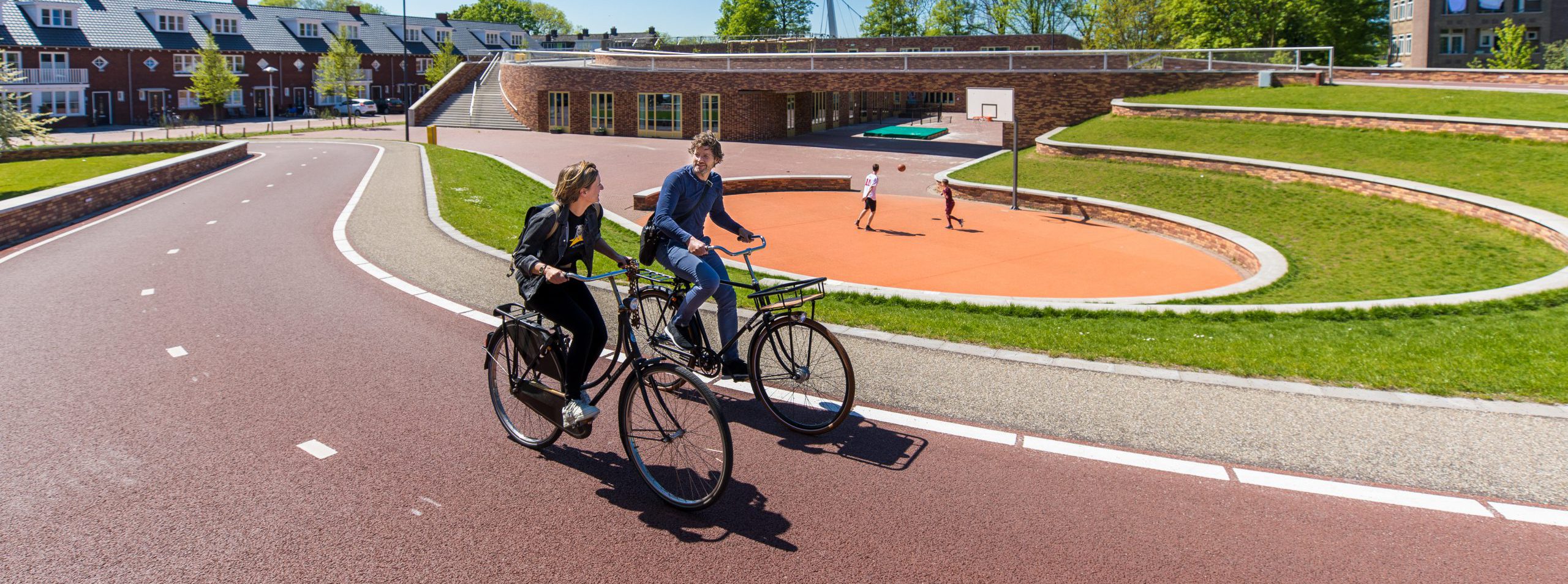 een rood kronkelend fietspad met twee fietsers. Op de achtergrond is een speelveld met gras in hoefijzer vorm in drie etages met in het midden een rond basketbalveld met oranje ondergrond. 