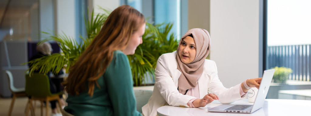 Twee vrouwelijke medewerkers zitten aan een tafel en kijken naar een laptop